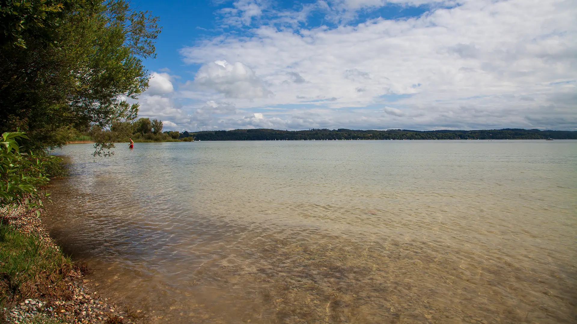 weiß blauer Himmel über dem Ammersee links das bewaldete Ufer und eine Person badet im Wasser. Am horizont sieht man das Ufer bis nach Herrsching am Ammersee
