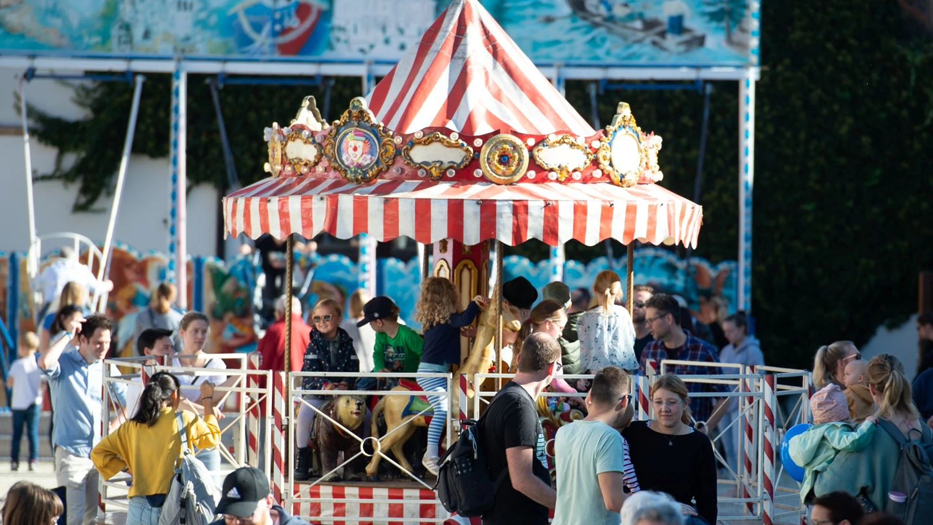 Ein nostalgisches Kinderkarussell mit rot-weiß gestreiftem Dach und vergoldeten Verzierungen steht im Sonnenlicht. Kinder sitzen auf den geschnitzten Tieren, rundherum stehen Besucherinnen und Besucher in Gruppen beisammen.