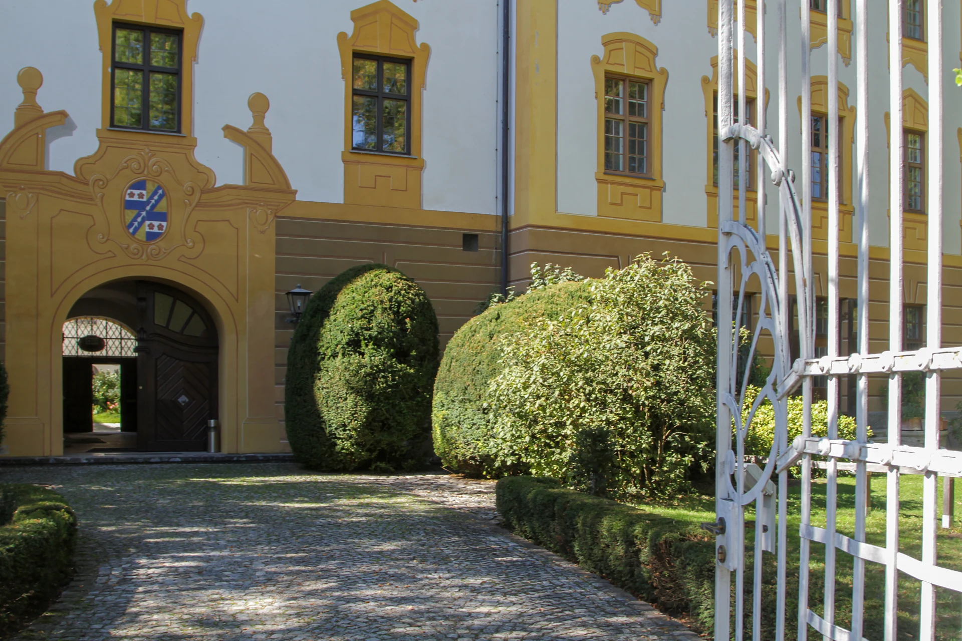 Blick in den Innenhof des Klosters Polling: eine gelb-weiße Barockfassade mit geschwungenen Giebeln, darunter ein Rundbogentor mit gemaltem Wappen und schwerer Holztür, davor kugelförmig geschnittene Buchsbäume, ein Kopfsteinpflasterweg und rechts ein weißes schmiedeeisernes Gartentor
