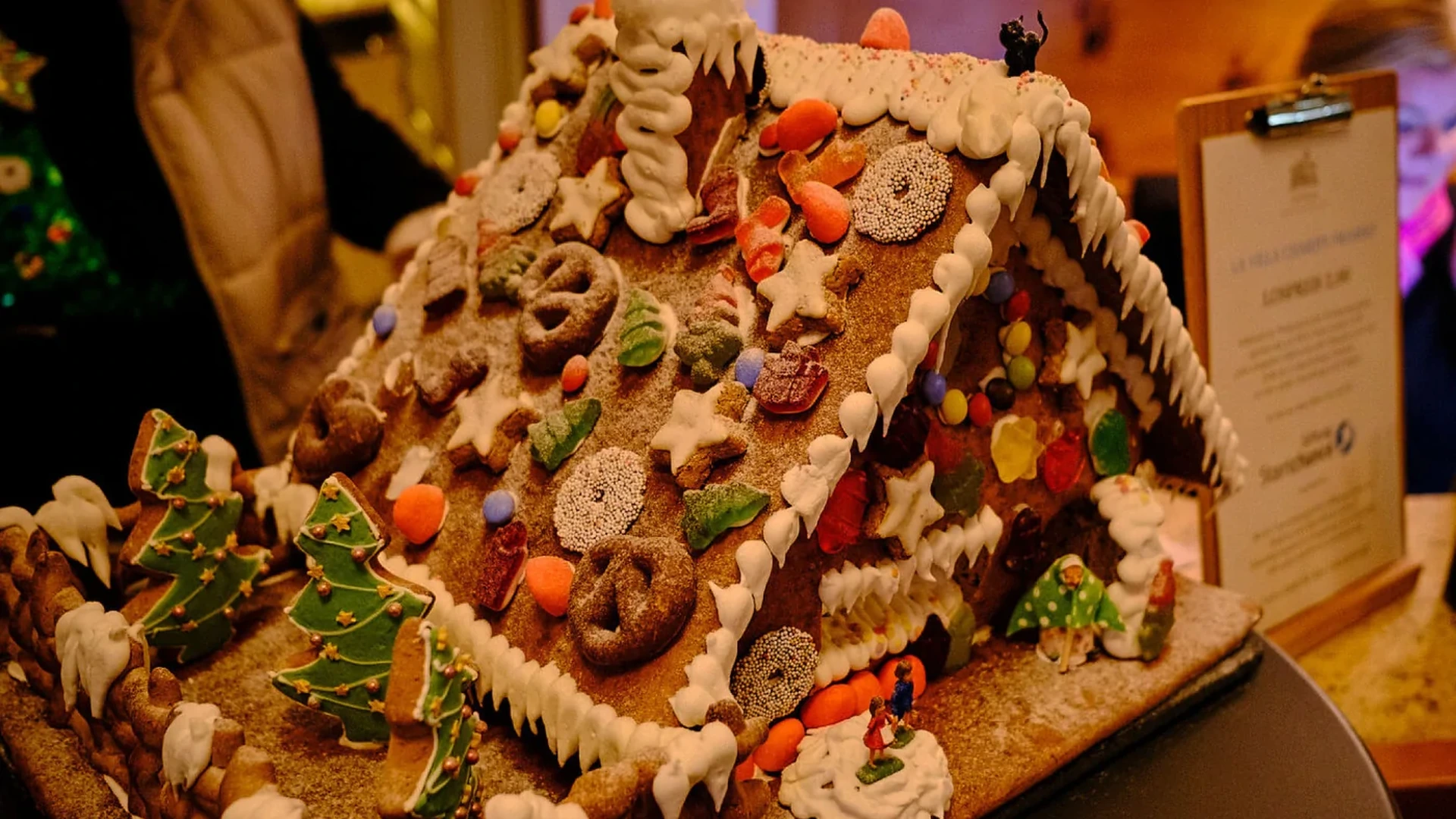 Ein reich verziertes Lebkuchenhaus auf einem Tablett. Dach und Wände sind mit weißem Zuckerguss, bunten Schokolinsen, Zuckerperlen, Brezeln und Sternen belegt, davor stehen grün verzierte Tannenbäume aus Teig und kleine Figuren im Schnee.