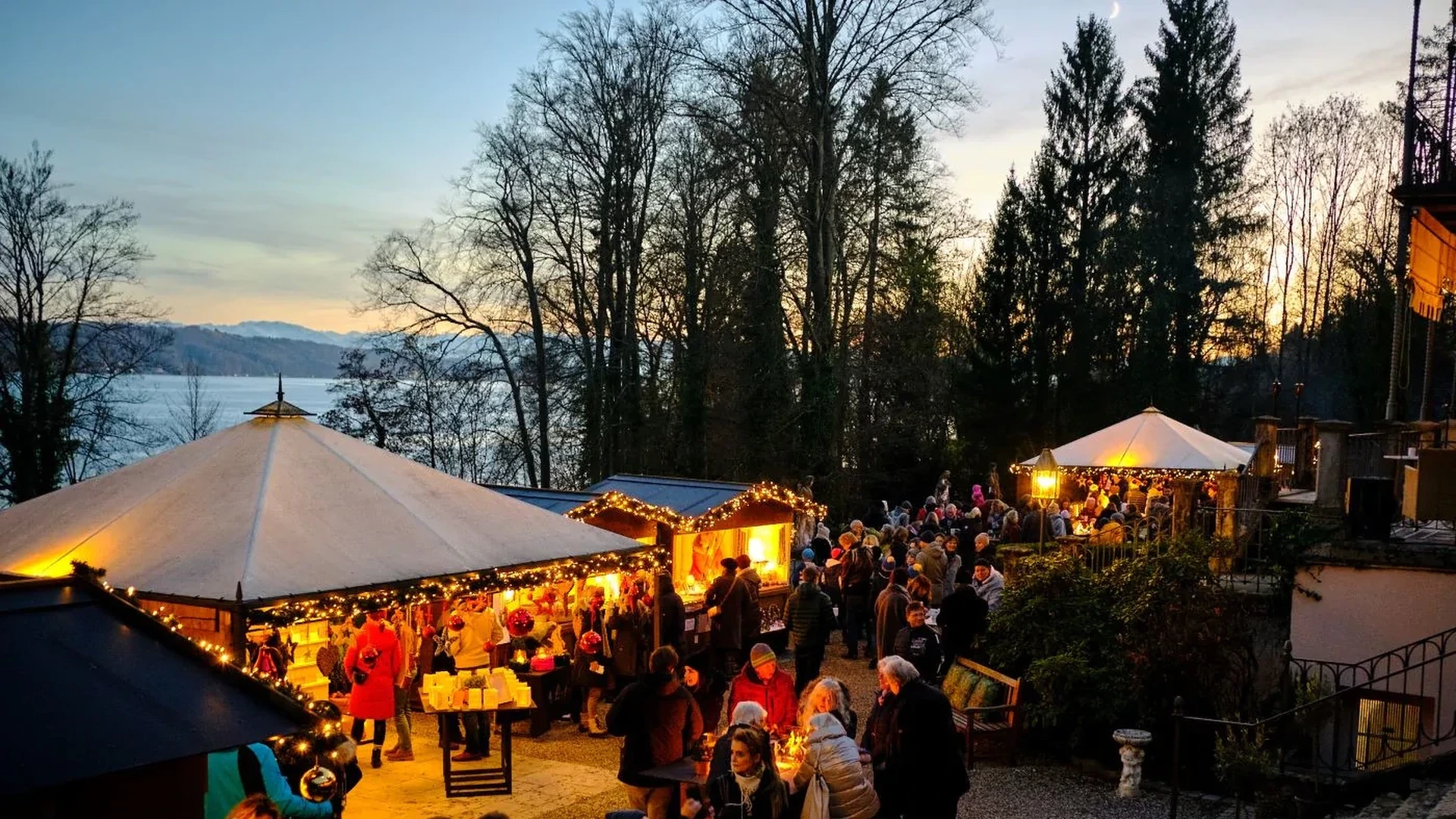 Abendlicher Weihnachtsmarkt im Park der LA VILLA am Starnberger See. Unter beleuchteten Pagodenzelten und Holzhütten mit Lichterketten stehen Besucherinnen und Besucher in Wintermänteln. Dahinter der See, kahle Bäume und die Alpenkette vor orange-blauem Abendhimmel.