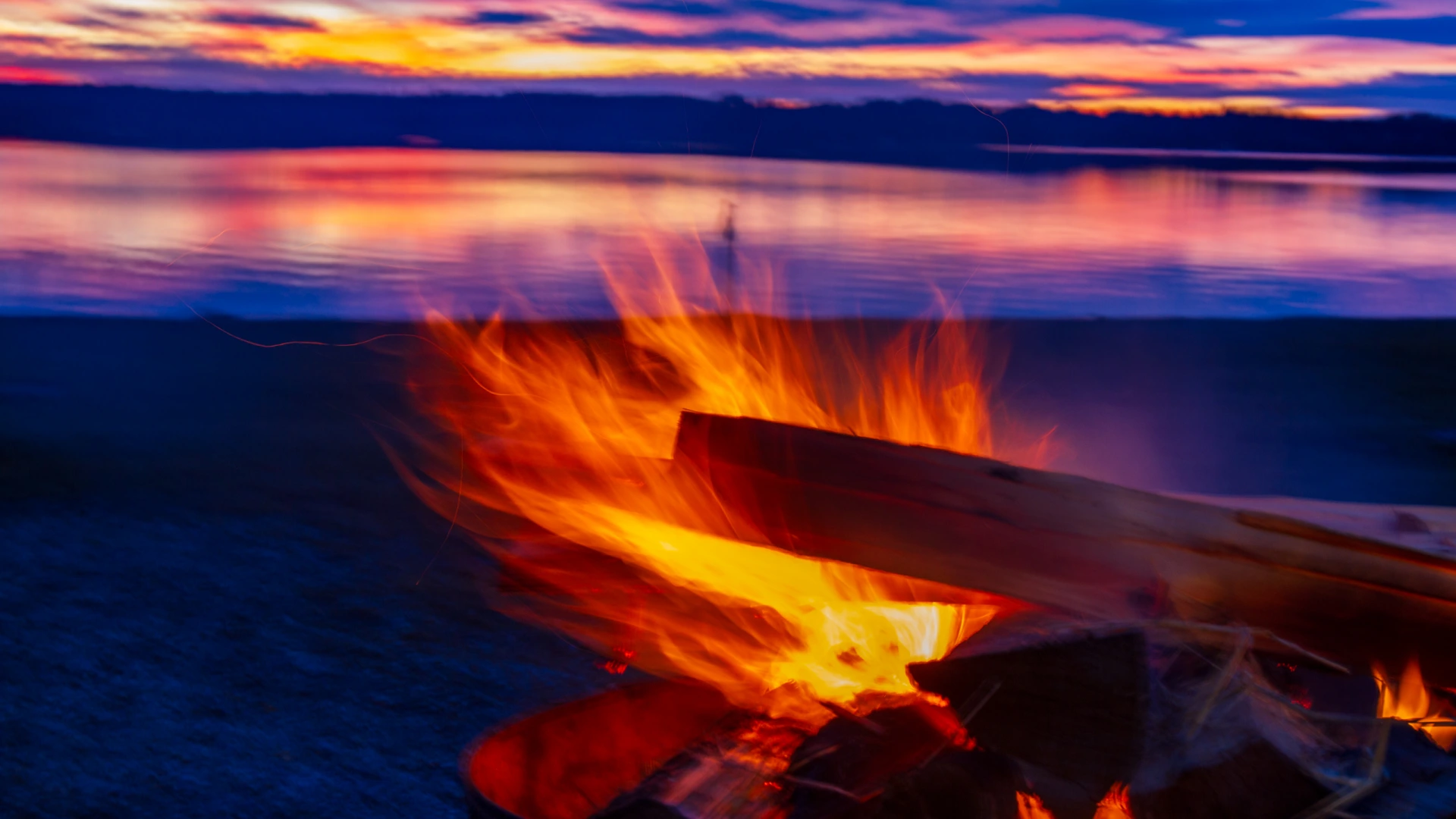 fuenfseen-lagerfeuer-starnberger-see-16-9-c-bwidmann Ein offenes Lagerfeuer am Kiesufer des Starnberger Sees. Die Flammen sind durch die lange Belichtungszeit zu einem gelb-orangen Strom verwischt, Funken ziehen helle Linien. Dahinter die ruhige Seefläche und ein violett-oranger Abendhimmel über der Uferlinie.