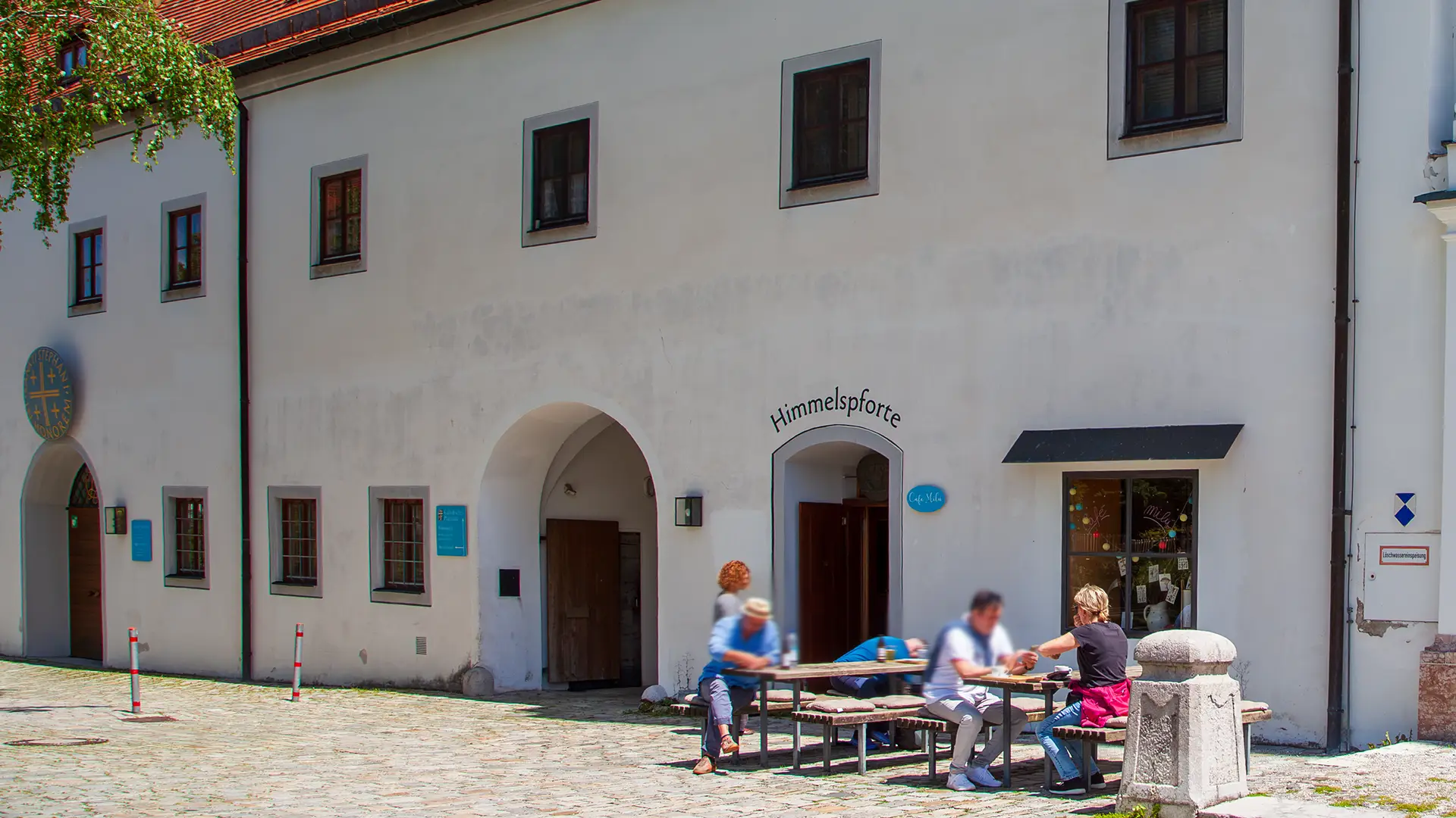 ein weißer historisches Gebäudewand mit Rundbögen und Fenstern davor auf Bierbänken ein paar Touristen vor dem Cafe - auf einem Rundbogen steht Himmelspforte.