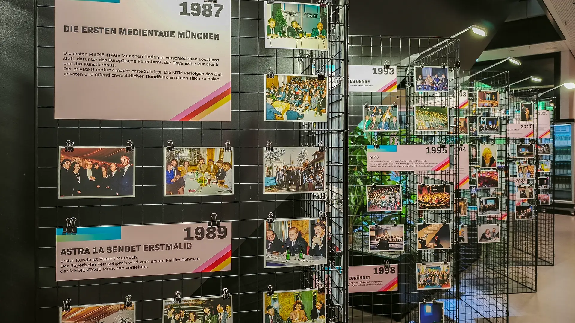 Fotogalerie auf Drahtzaungestellen mit Fotos und Schautafeln von 1987 bis 2026 - Highlights der Jahre wie Astra 1a sendet erstmalig 1987 die ersten Medientage München - gefolgt von 1993 1995 1998 und weiteren Schautafeln im Gang der Blue Stage.