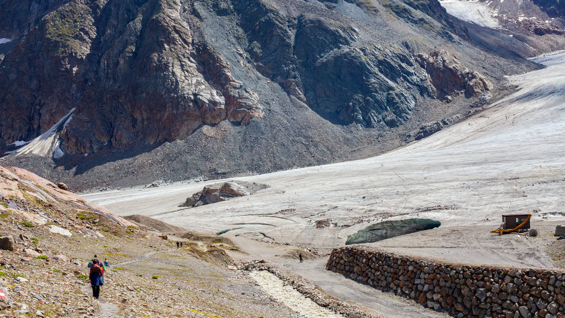 Blick über ein weites Geröllfeld auf die ausgeaperte Zunge des Pitztaler Gletschers, rechts eine Pistenraupe und eine Steinmauer am Gletscherrand, darüber dunkle Felswände.