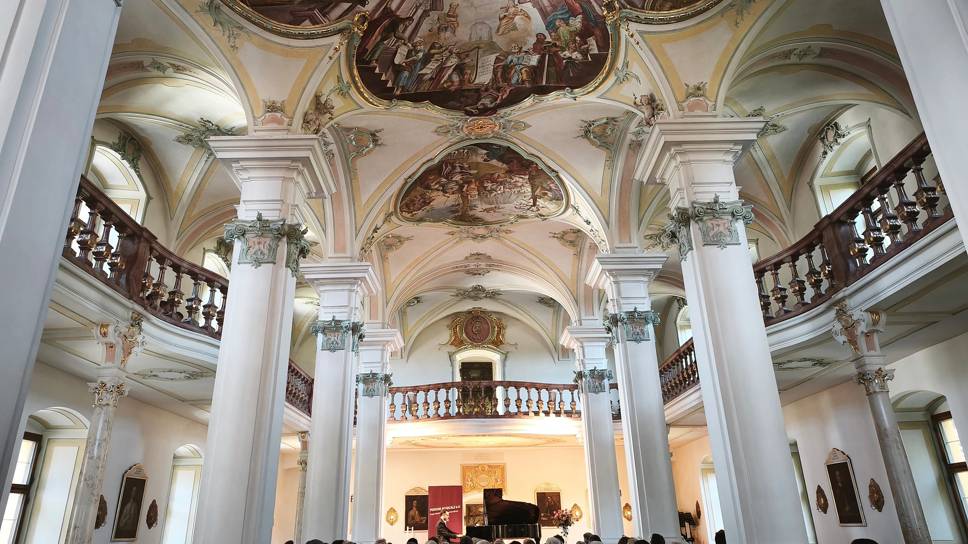 Blick in den barocken Bibliothekssaal des Klosters Polling mit Deckenfresken, weißen Säulen und umlaufender Empore; vorne spielt der Pianist Johannes Obermeier am Flügel vor vollbesetzten Stuhlreihen, daneben ein rotes Banner mit dem Schriftzug Podium musicale e.V.
