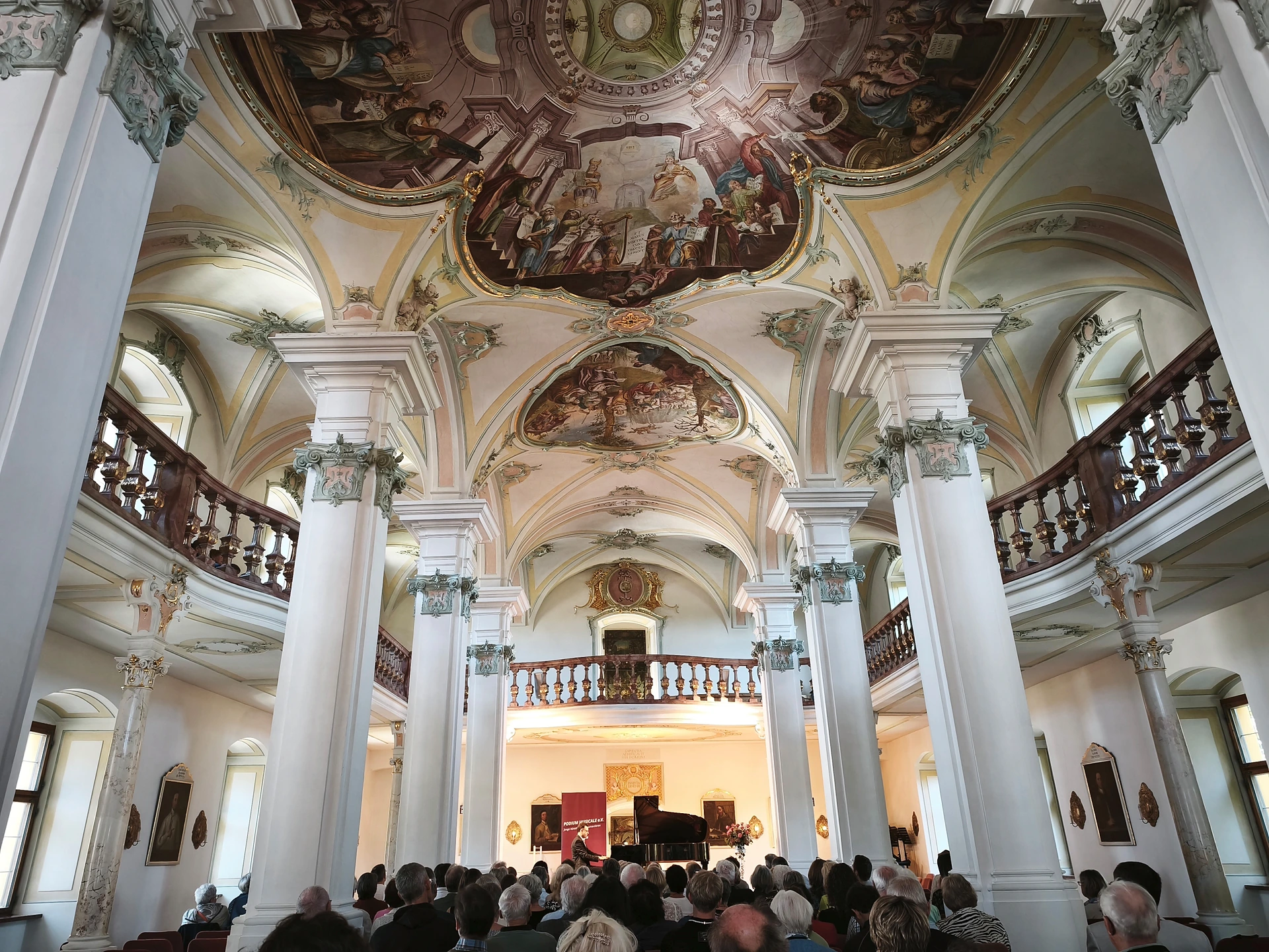 Blick in den barocken Bibliothekssaal des Klosters Polling mit Deckenfresken, weißen Säulen und umlaufender Empore; vorne spielt der Pianist Johannes Obermeier am Flügel vor vollbesetzten Stuhlreihen, daneben ein rotes Banner mit dem Schriftzug Podium musicale e.V.