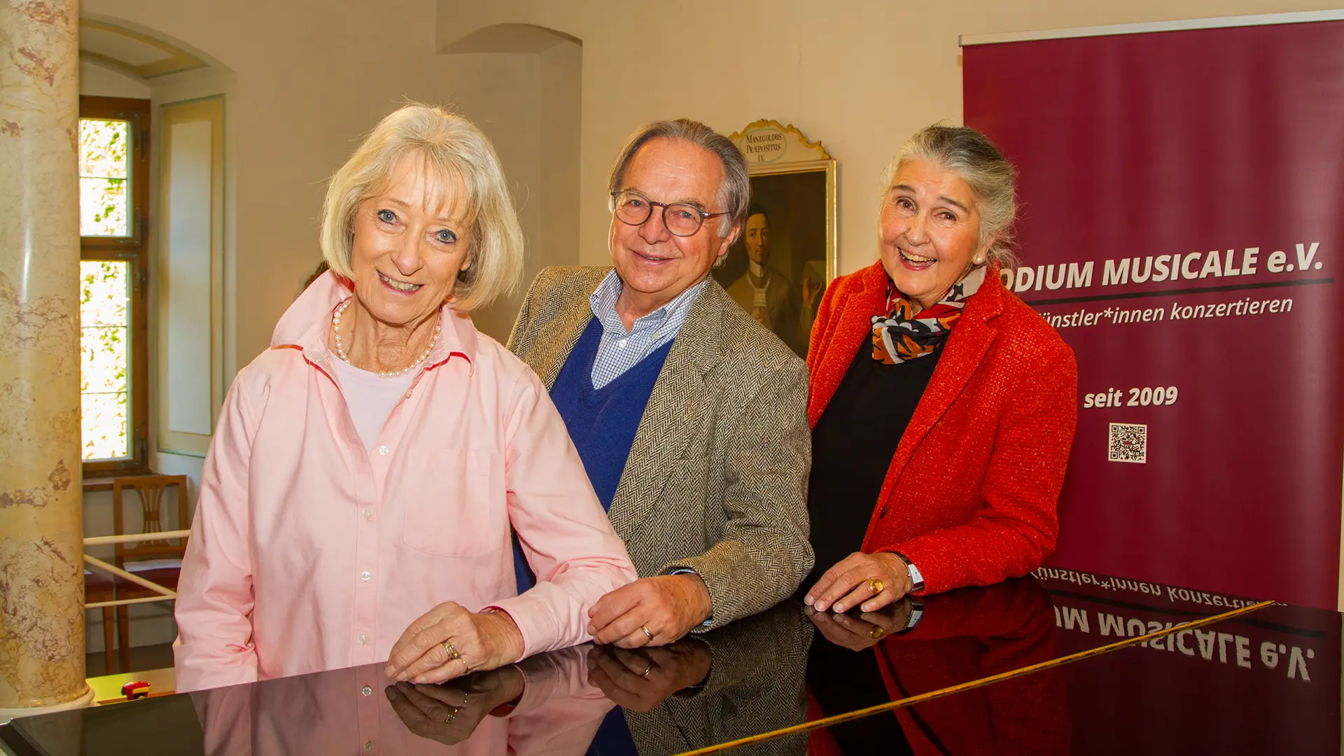 antiker Saal mit dem Plakat Podium musicale e.v. eine frau in rosa Bluse, ein Mann mit Brille und brauenen Sakko und eine Frau in rotem Jacket lehnen auf einem Fllügel und lächeln in die Kamera