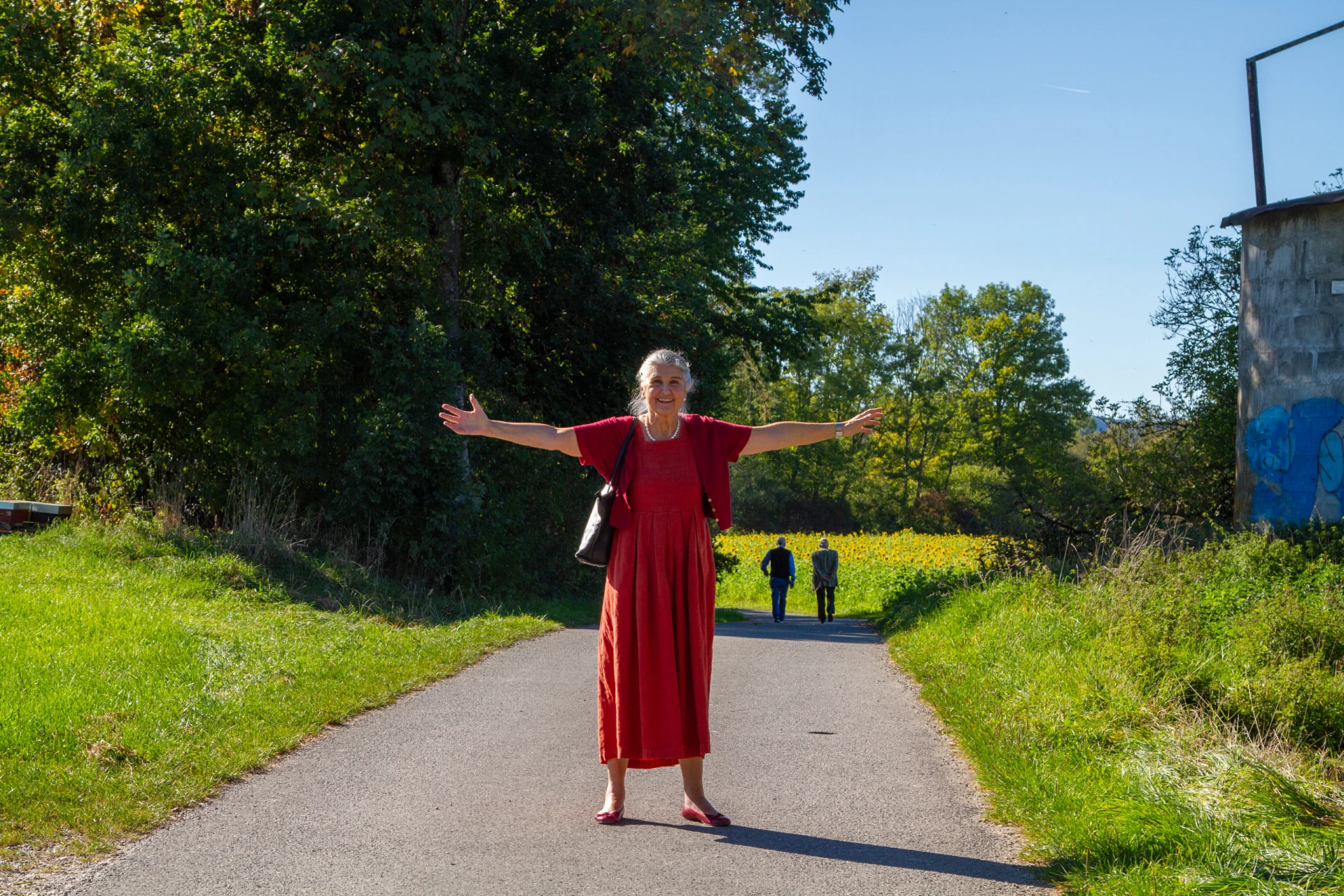 Ursula Hauer steht in rotem Kleid mit weit ausgebreiteten Armen lachend auf einem schmalen asphaltierten Feldweg, links hohe Bäume, rechts ein blühendes Sonnenblumenfeld, im Hintergrund gehen zwei Personen den Weg entlang, strahlend blauer Himmel