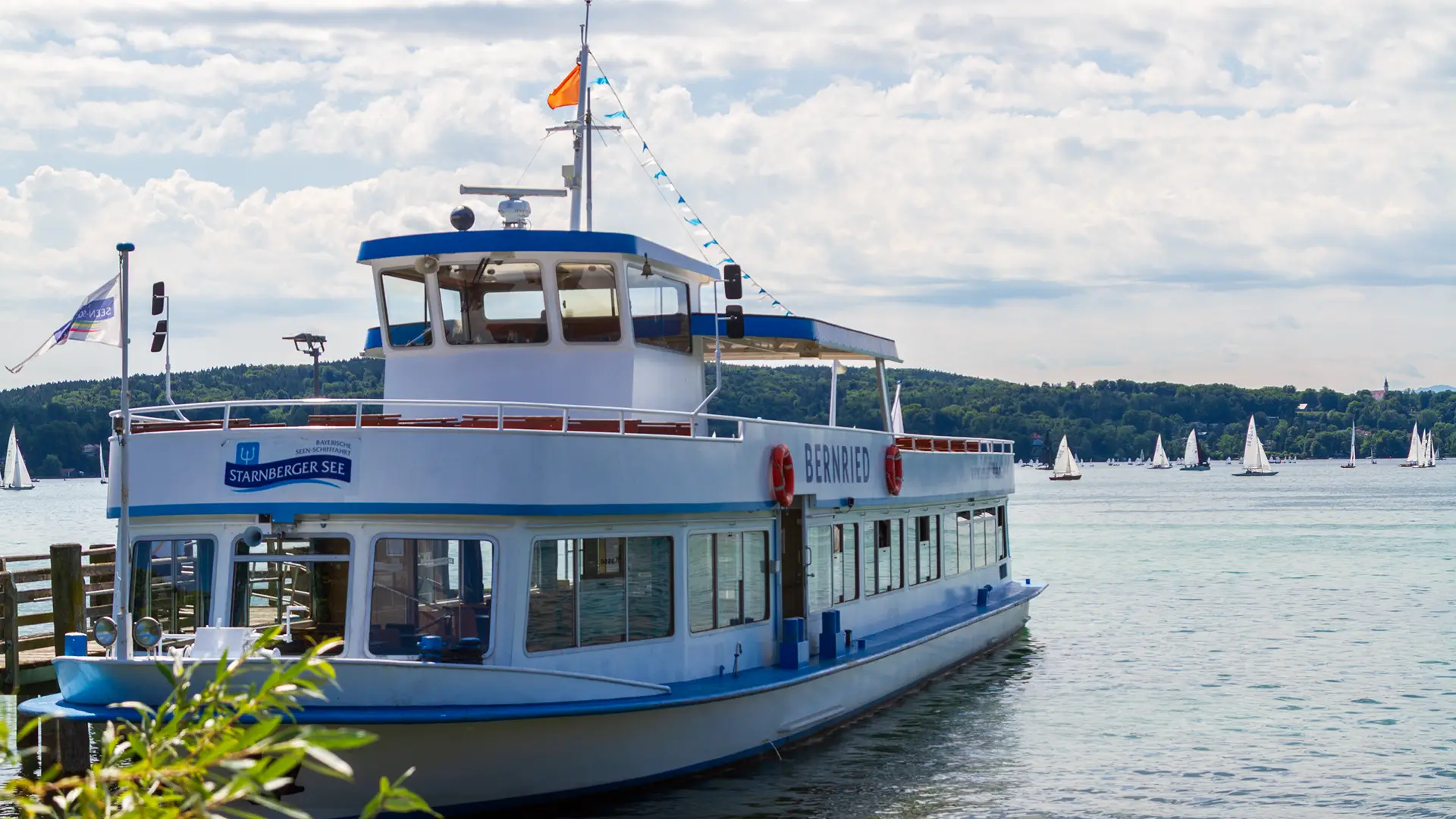 MS Bernried der Bayerischen Seenschifffahrt Starnberger See liegt am Landungssteg in Tutzhing im Hintergrund Segelschiffe und das bewaldete Ufer bei wolkigen Himmel.