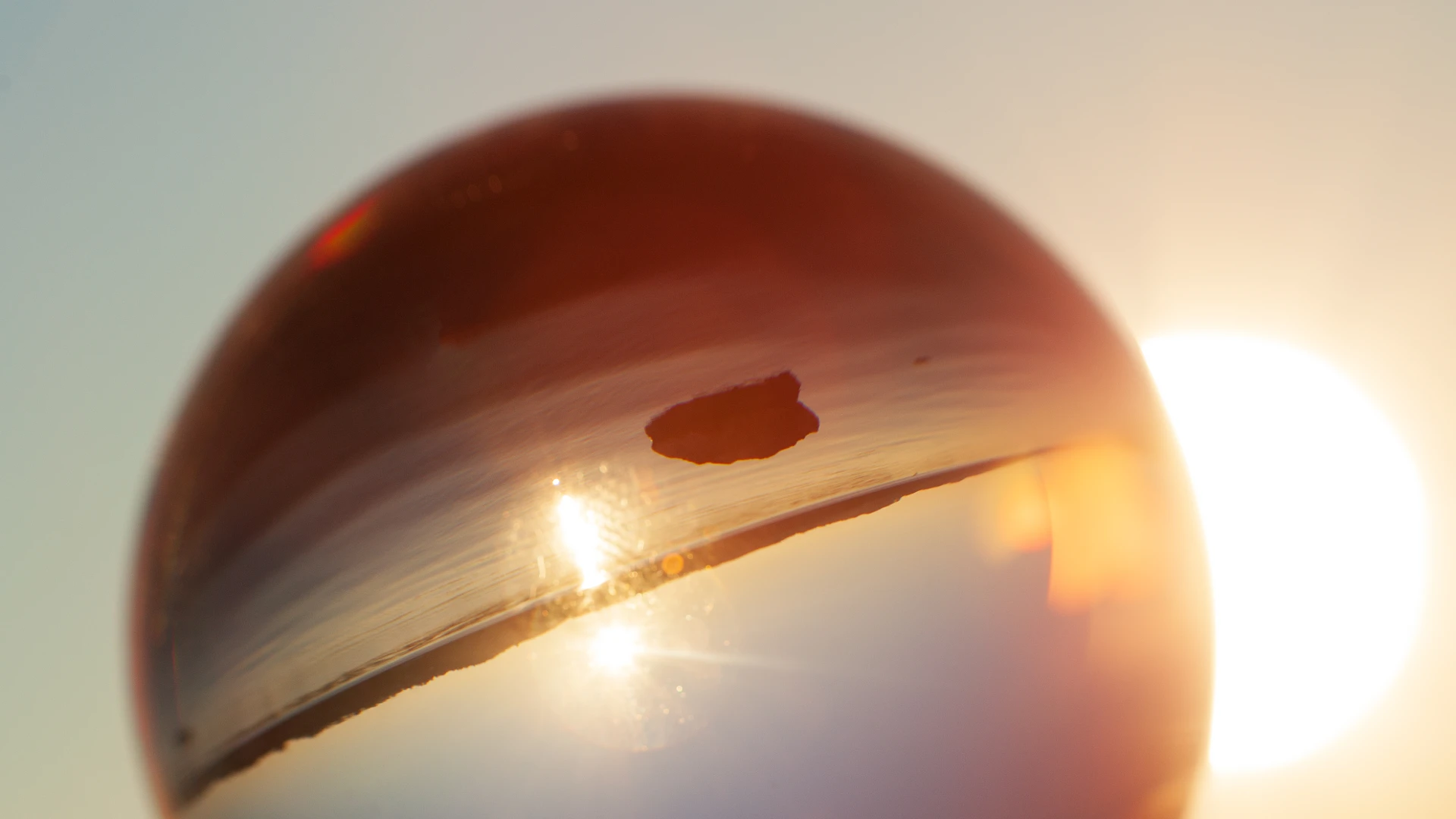 Eine gläserne Kugel vor der tiefstehenden Sonne über dem Starnberger See. In der Kugel erscheint die Landschaft auf dem Kopf: Die Uferlinie und der glitzernde Sonnenstreifen auf dem Wasser stehen kopfüber im Glas, dahinter geht die echte Sonne unter.