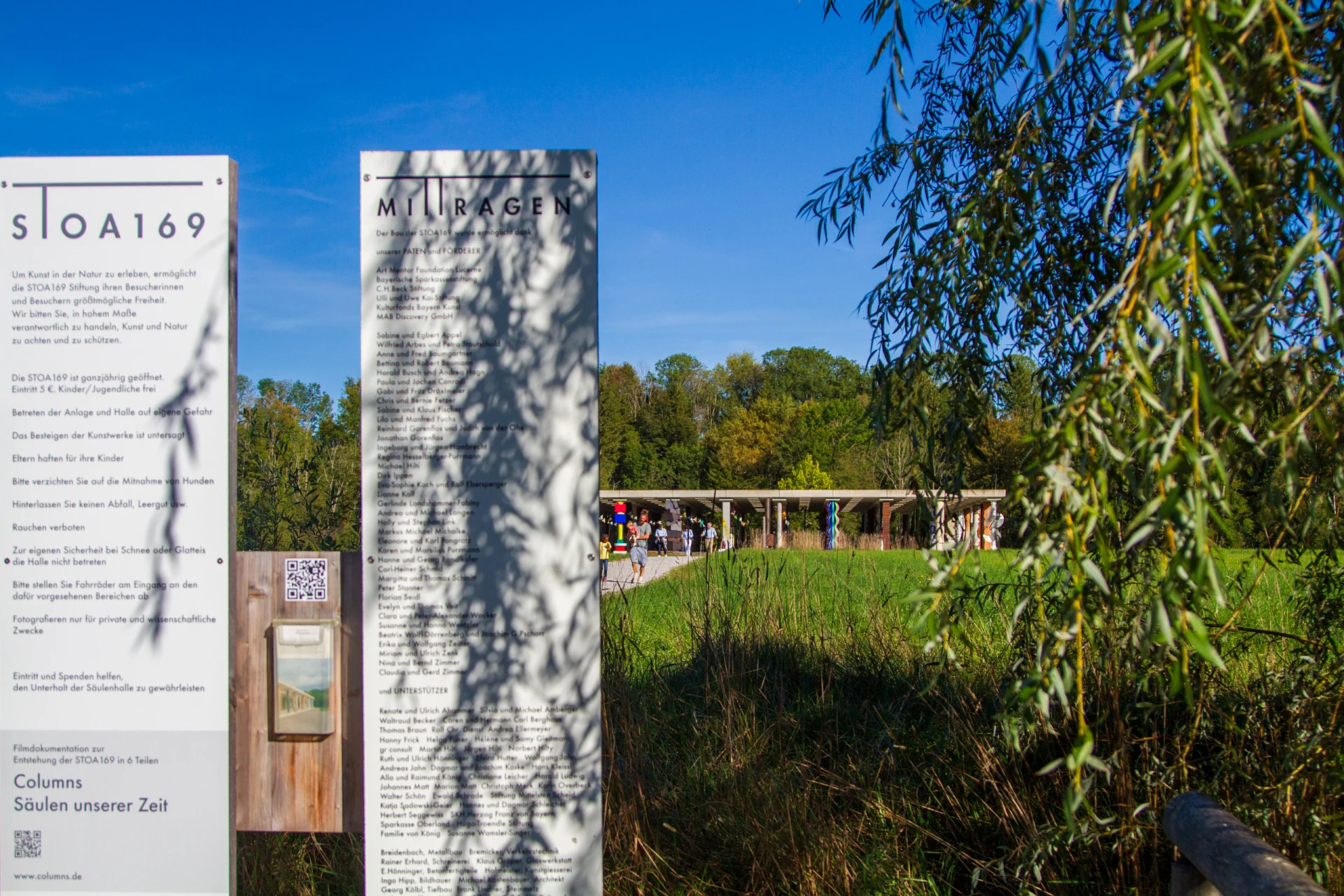 Zwei weiße Infotafeln am Eingang der Stoa 169 mit Besucherhinweisen und einer Liste der Förderer, dahinter über einer Wiese die offene Säulenhalle mit bunten Säulen, rechts hängende Weidenzweige, blauer Himmel