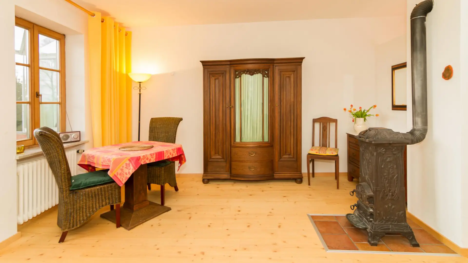 Wohnraum Ferienwohnung Seeblick mit Holzfußboden, antikem Holzofen, antikem Schrank, Sitzensemble vor dem Fenster im Tageslicht.