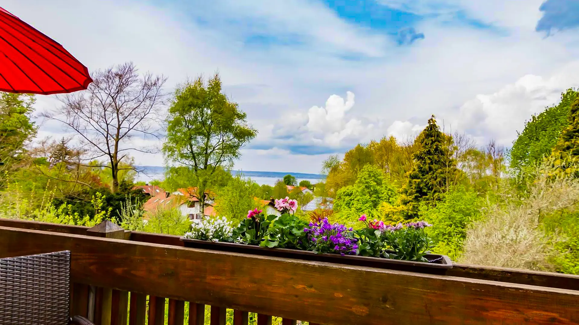 Blick vom Balkon der Ferienwohnung Seeblick auf den Ammersee bei weiß blauem Himmel und Sonnenschein. im Vordergrund ein Sonnenschirm und Balkonpflanzen.