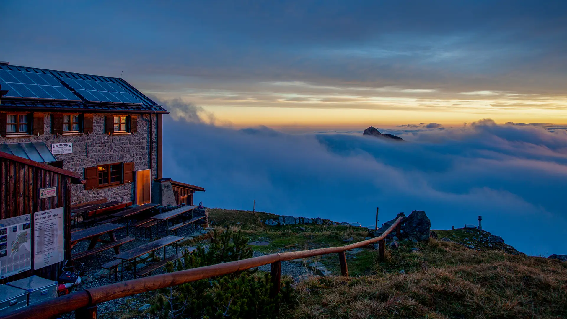 Weilheimer Hütte - steinerne Berghütte im warmen Licht des Sonnenaufgangs, rechts daneben ein Meer aus Wolken, aus dem eine einzelne Felsspitze ragt; der Himmel geht von Orange in tiefes Blau über.