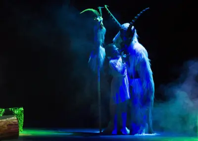 Blau angestrahlte zottelige Gestalten mit langen gebogenen Hörnern stehen im Bühnennebel. Zwischen ihnen eine hell gekleidete Figur.