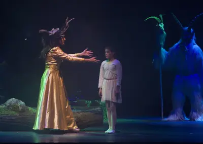 Die Frau im goldenen Kleid breitet beide Arme aus, ihr gegenüber steht das Mädchen in Weiß. Links und rechts ragen blaue Perchten ins Bild.