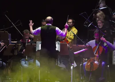 Ein Dirigent mit erhobenen Händen steht vor dem Orchester. Streicher, ein Cello und Notenpulte sind zu erkennen.