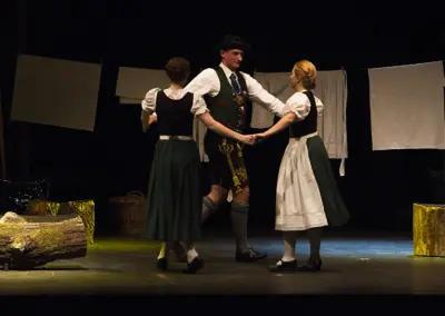 Ein Mann in Lederhose tanzt mit zwei Frauen in Dirndl. Im Hintergrund hängen weiße Tücher an Wäscheleinen, links steht ein Weidenkorb.