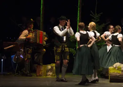 Eine Gruppe in Tracht tanzt in Reihe, Männer in Lederhosen und Frauen in grünen Dirndlröcken. Links spielen Musiker mit Akkordeon.