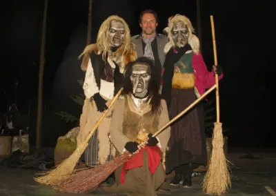 Drei Figuren mit geschnitzten Hexenmasken, Perücken und Besen stehen beieinander. Zwischen ihnen ein Mann im Anzug mit Schal, der in die Kamera blickt.